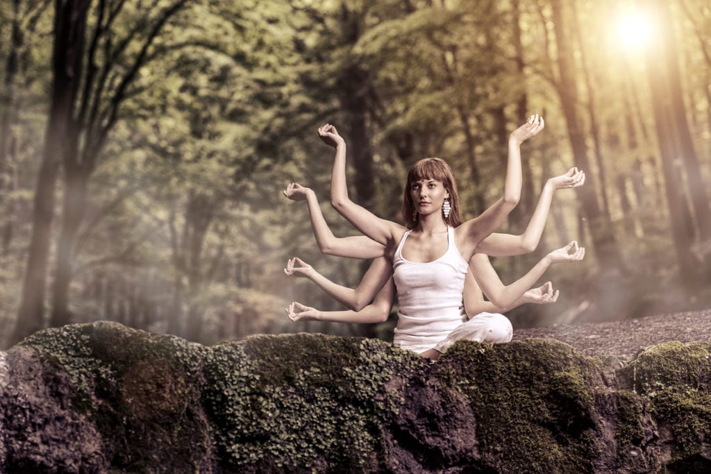 A young woman meditates in a forest, the sun filters through the trees. Her eight arms are in a pose like goddess Kali.